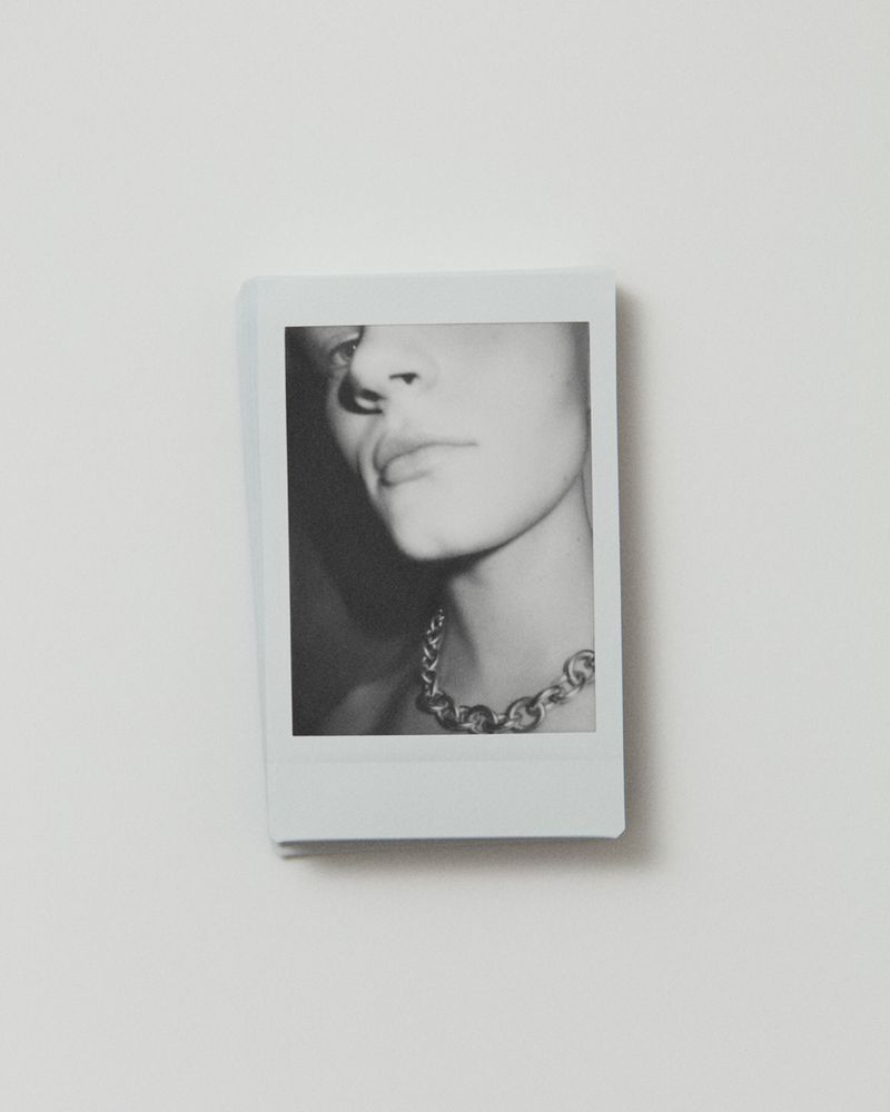 Black and white photograph of a young adult with a metal chain necklace, indoor setting, shallow depth of field, clean presentation.
