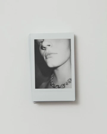 Black and white photograph of a young adult with a metal chain necklace, indoor setting, shallow depth of field, clean presentation.