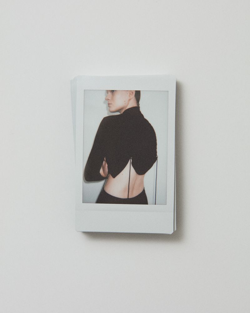Polaroid image featuring a woman in a black top with an open back, arms crossed, and looking to the side against a white background.
