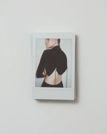 Polaroid image featuring a woman in a black top with an open back, arms crossed, and looking to the side against a white background.