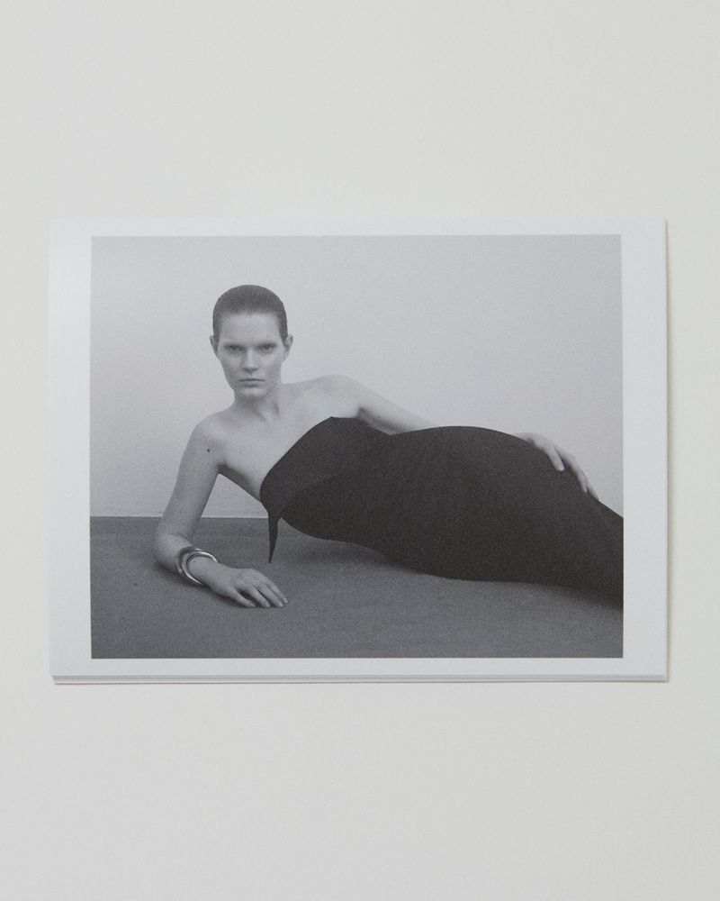 Monochrome fashion shot of a woman in a strapless black dress reclining against a grey surface against a plain, light-toned background.