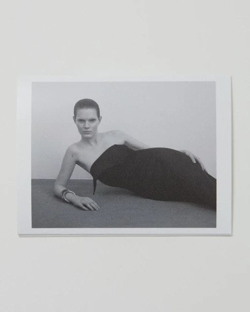 Monochrome fashion shot of a woman in a strapless black dress reclining against a grey surface against a plain, light-toned background.