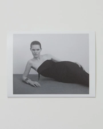 Monochrome fashion shot of a woman in a strapless black dress reclining against a grey surface against a plain, light-toned background.