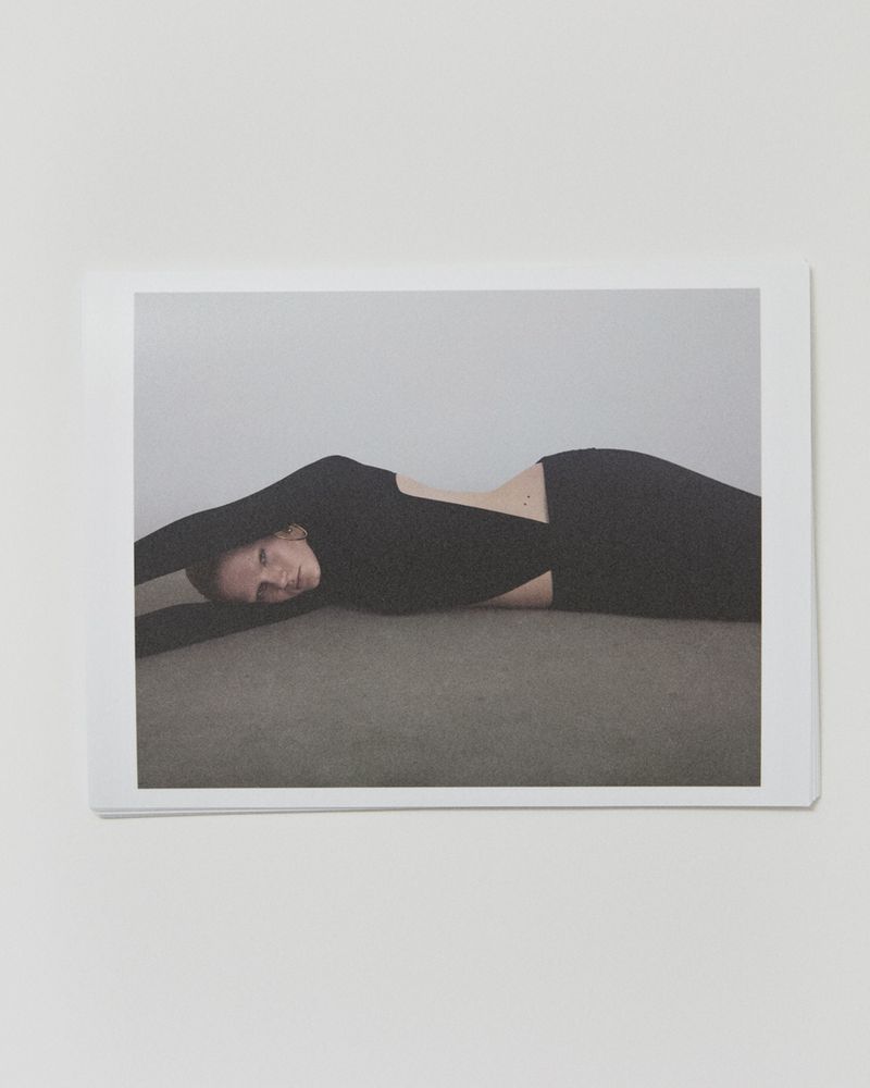 Woman with black long-sleeve dress lying on gray surface in a minimalist studio, arms extended, captured in a fashion-style indoor portrait.