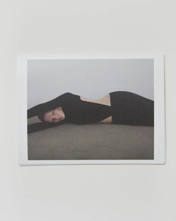 Woman with black long-sleeve dress lying on gray surface in a minimalist studio, arms extended, captured in a fashion-style indoor portrait.