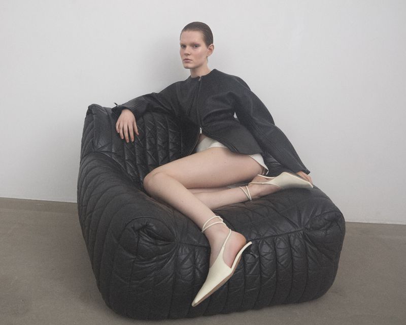 Model wearing dark jacket sitting on a large black quilted armchair against a white background