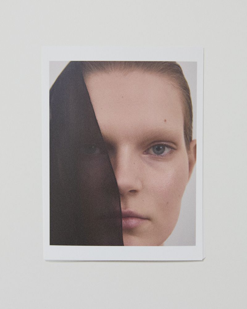 Close-up portrait of a model with a dark sheer fabric draped partially over her face against a plain background.