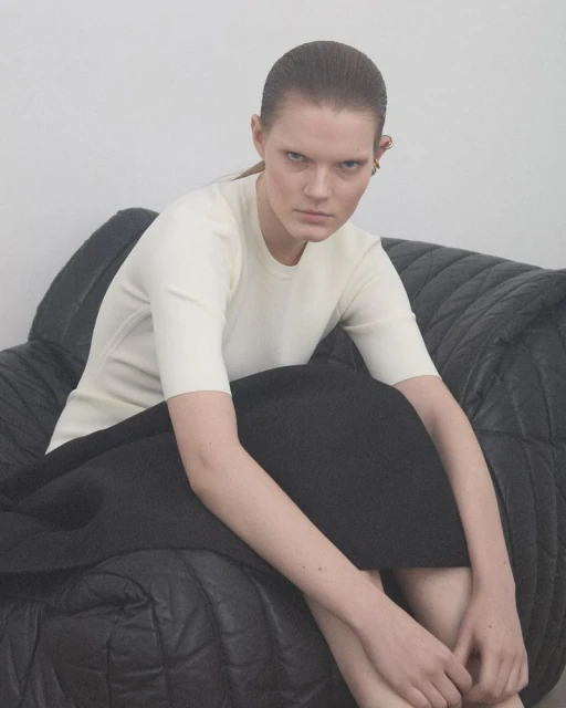 Adult woman with slicked-back hair, wearing a cream-colored shirt and black skirt, seated on a quilted black sofa in a studio setting.