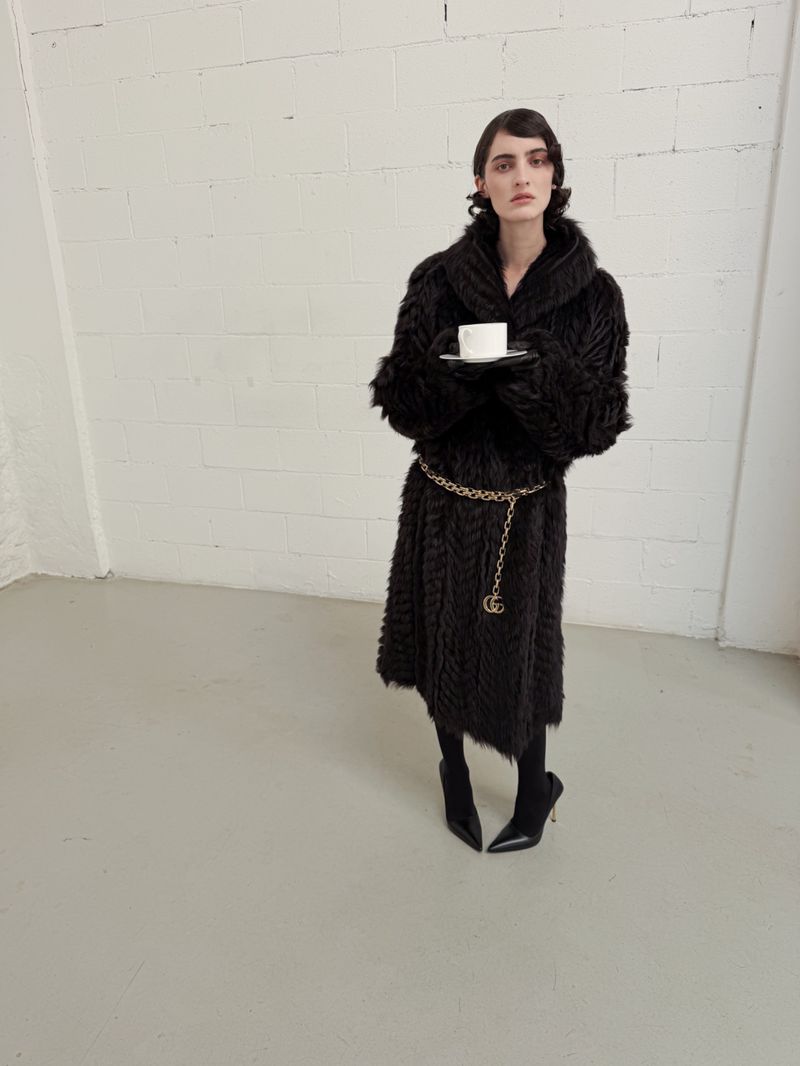 A woman in a black textured coat and heeled boots stands in a white studio holding a cup and saucer in a fashion editorial shot.
