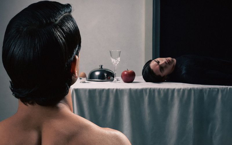 Rear view of a dark-haired man facing a table with an egg, covered platter, glass, apple, and another dark-haired person laying on the table.