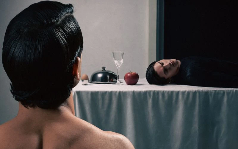Rear view of a dark-haired man facing a table with an egg, covered platter, glass, apple, and another dark-haired person laying on the table.