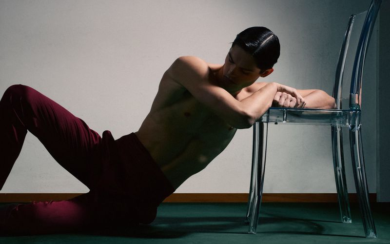 Man lying down shirtless in red trousers leaning on clear plastic chair in a studio with shadow and modern aesthetic. 