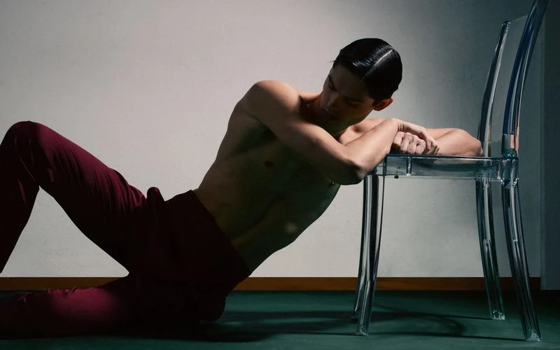 Man lying down shirtless in red trousers leaning on clear plastic chair in a studio with shadow and modern aesthetic. 