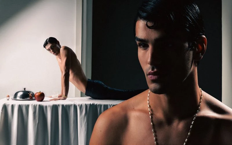 Stylish studio shot of two shirtless men with dark hair in front of an egg, apple, and covered platter on a white table.