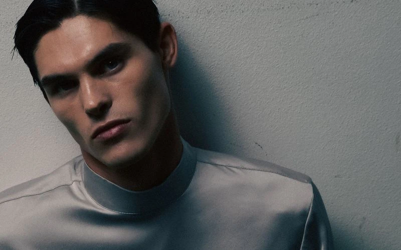 Headshot of a fair-skinned adult male with blue eyes, dark hair neatly styled, wearing a light grey high-necked satin shirt against a grey wall.
