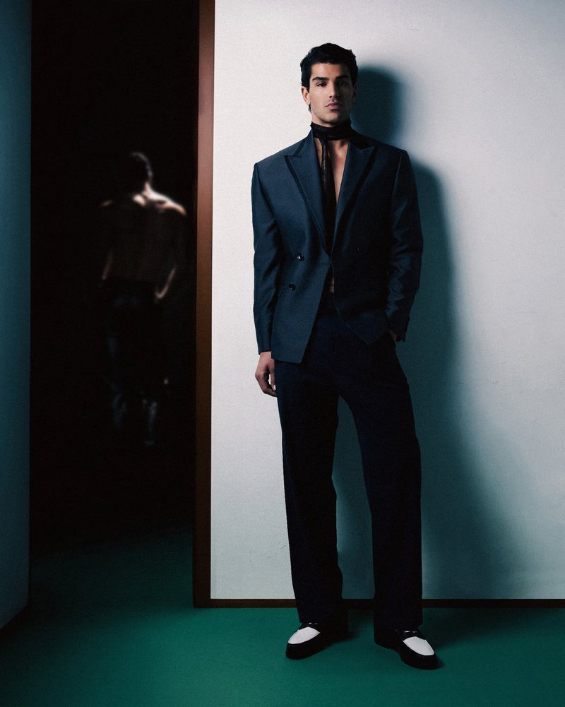 Full-length shot of a handsome man in a dark suit with a scarf, posing in a modern interior with a glimpse of a second person in the background.