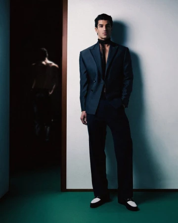 A man wearing a dark navy double-breasted suit and a black neck scarf stands against a white wall next to a dark doorway.