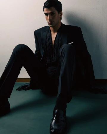 Young man in a black tuxedo and pinstriped vest sitting on the floor against a plain wall looking at the camera.