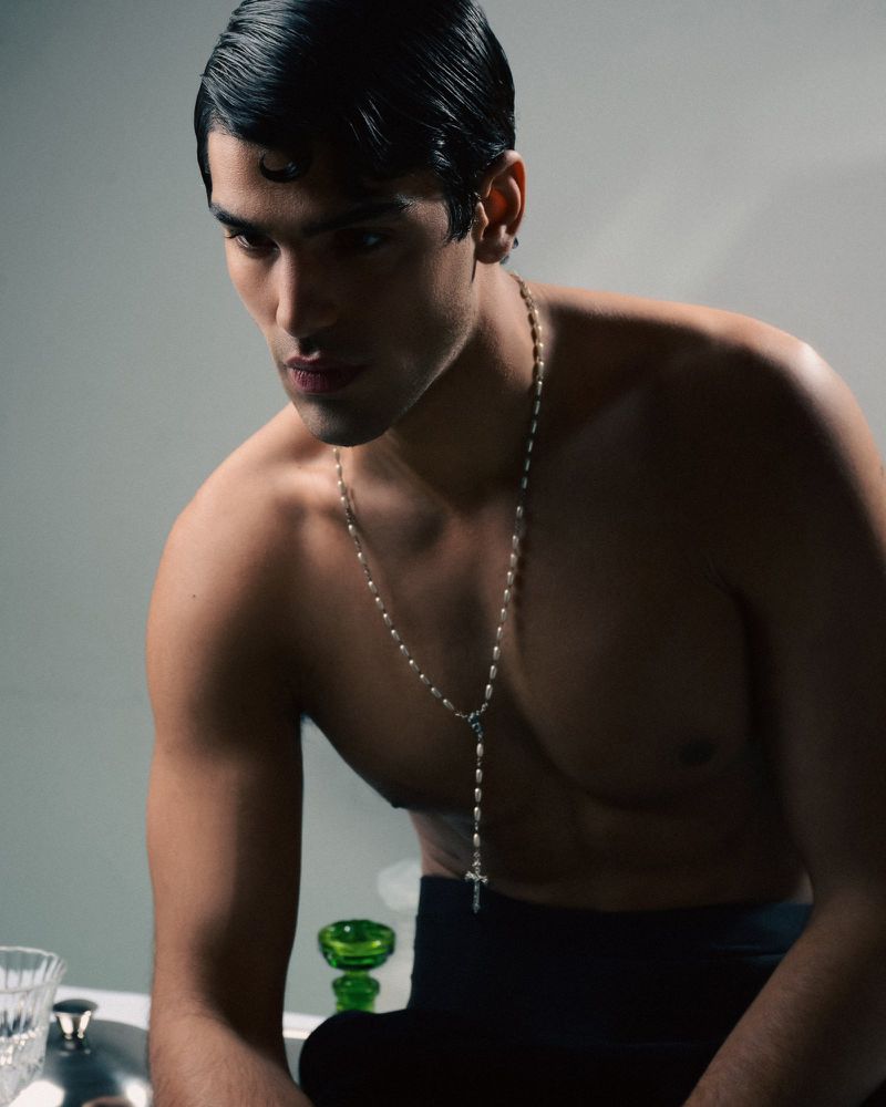 Portrait of shirtless adult male with slicked-back dark hair, wearing a silver rosary necklace, posing in a neutral lit indoor setting.