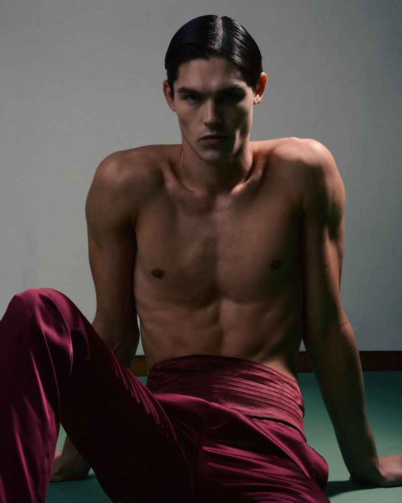 Portrait of a male fashion model with a serious expression. He is shirtless wearing red pants, in a studio with neutral lighting.