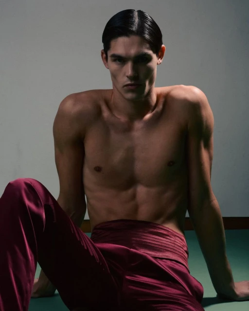 Portrait of a male fashion model with a serious expression. He is shirtless wearing red pants, in a studio with neutral lighting.