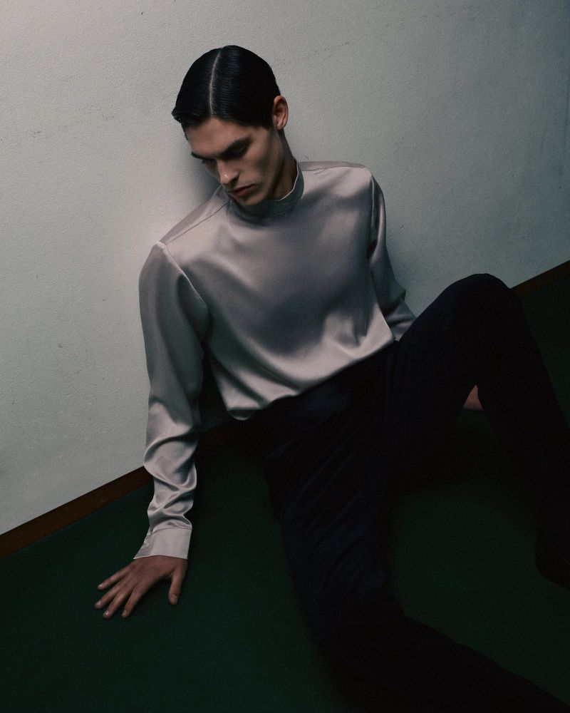 A young adult male model poses indoors wearing a silver satin long-sleeve shirt and black pants, leaning against a white wall with a green floor.