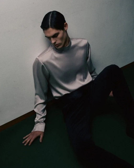 A young adult male model poses indoors wearing a silver satin long-sleeve shirt and black pants, leaning against a white wall with a green floor.