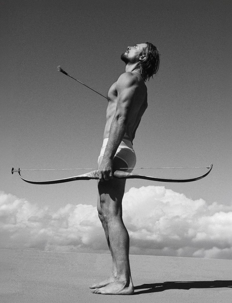 Gabriel Aubry standing outdoors holding a long bow with an arrow piercing his chest against a sky and horizon background