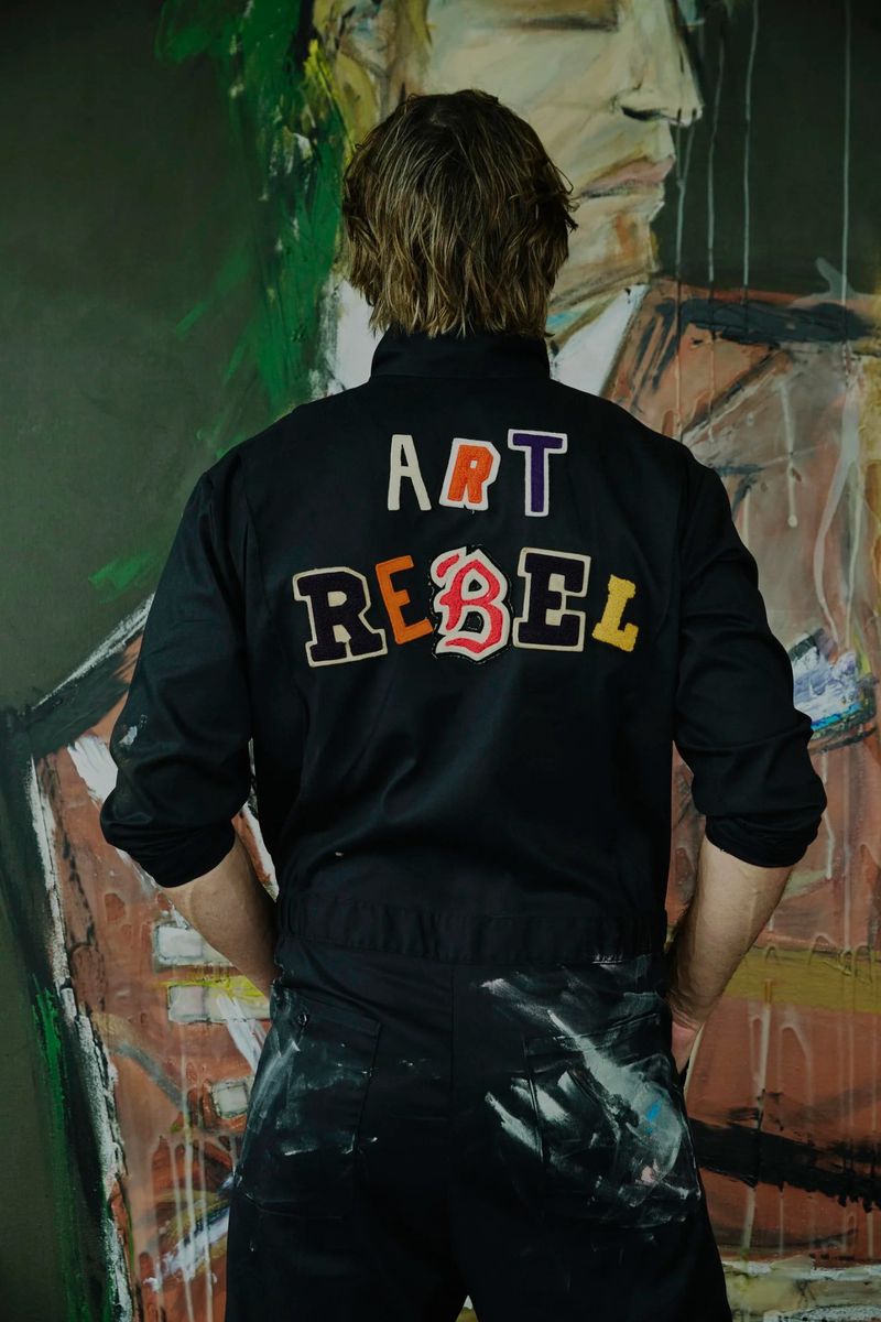 Back view of a fair-skinned adult wearing black overalls with the text 'Art Rebel' on the back, standing in front of an art painting.