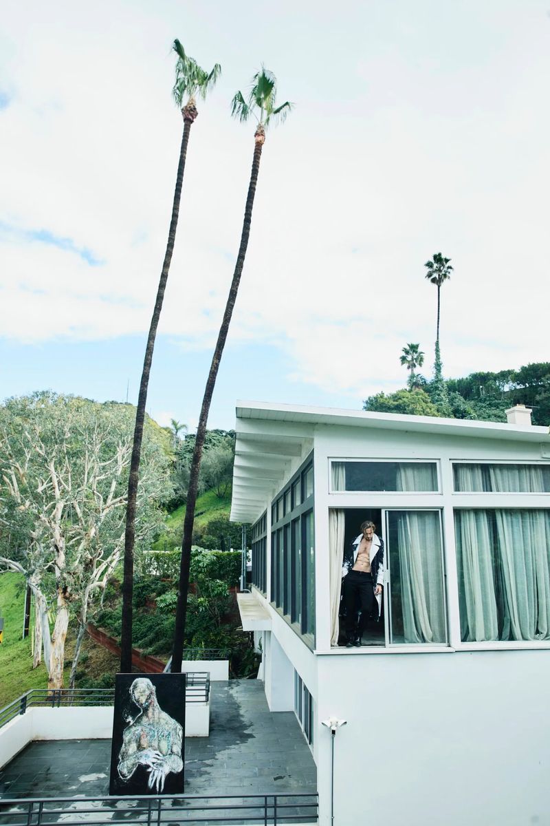 Fashionable adult male poses on a modern white balcony with palm trees. Artwork and lush green foliage accentuate the outdoor scene.