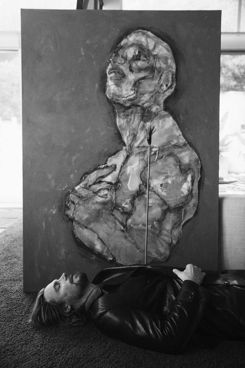 Black and white portrait of a man lying on the floor, next to a large painting of a human bust, in an indoor environment.