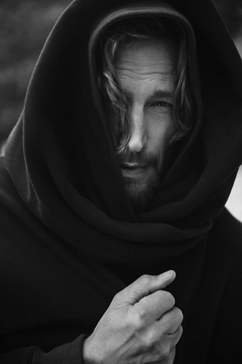Monochrome portrait of a man with long hair, partially obscured by a dark hooded shawl, standing outdoors with a neutral expression.