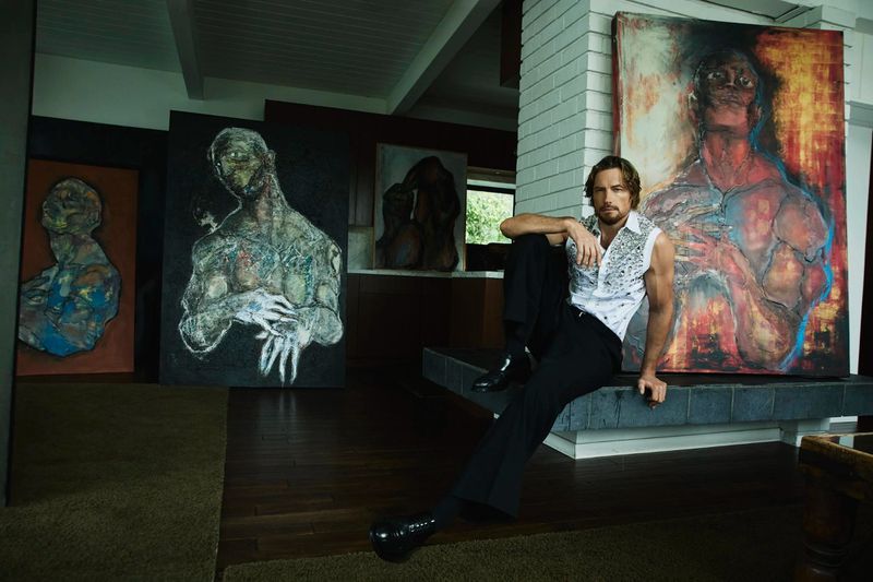Adult male artist poses next to his large paintings in a home setting, featuring natural light, interior brick wall, and creative atmosphere.