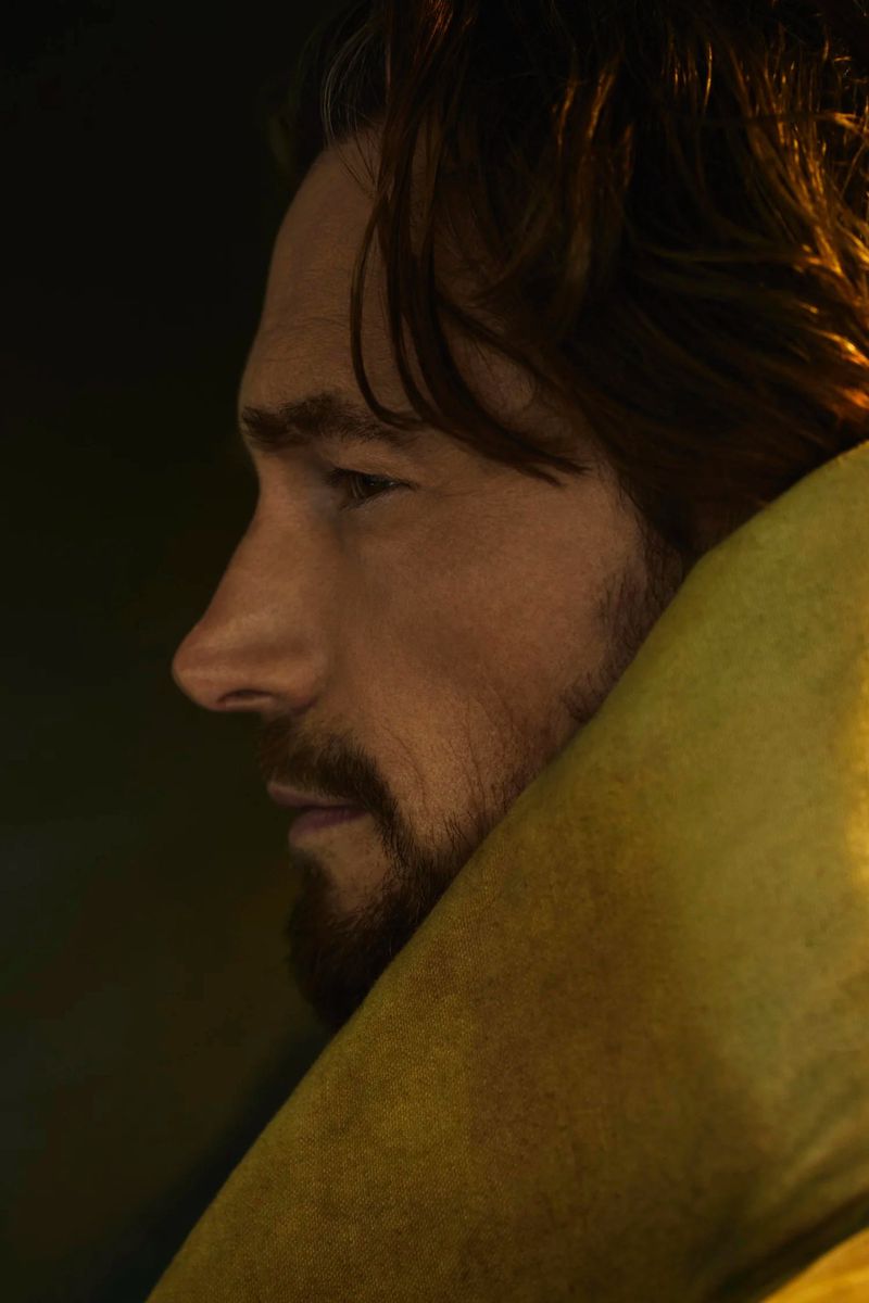 Profile portrait of an adult male with a beard and medium-length hair, facing left, with dramatic lighting and yellow fabric.