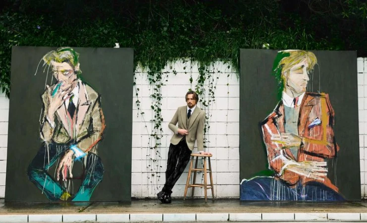A man in a suit leans against a stool between two portrait paintings in an outdoor setting with green foliage in the background.