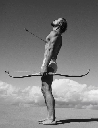 Gabriel Aubry standing outdoors holding a long bow with an arrow piercing his chest against a sky and horizon background