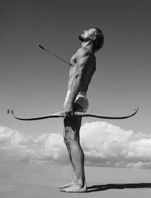 Gabriel Aubry standing outdoors holding a long bow with an arrow piercing his chest against a sky and horizon background