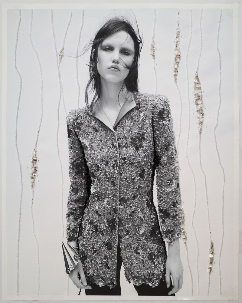 Monochrome portrait of a slender adult woman posing in a textured blazer with wind-blown dark hair against a patterned backdrop.
