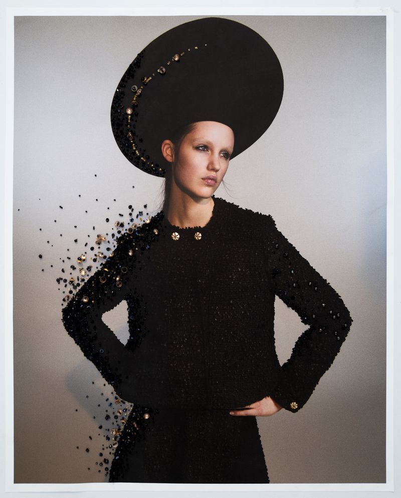 Woman in a black embellished jacket and a large brimmed hat stands against a light background in a fashion editorial style photograph.