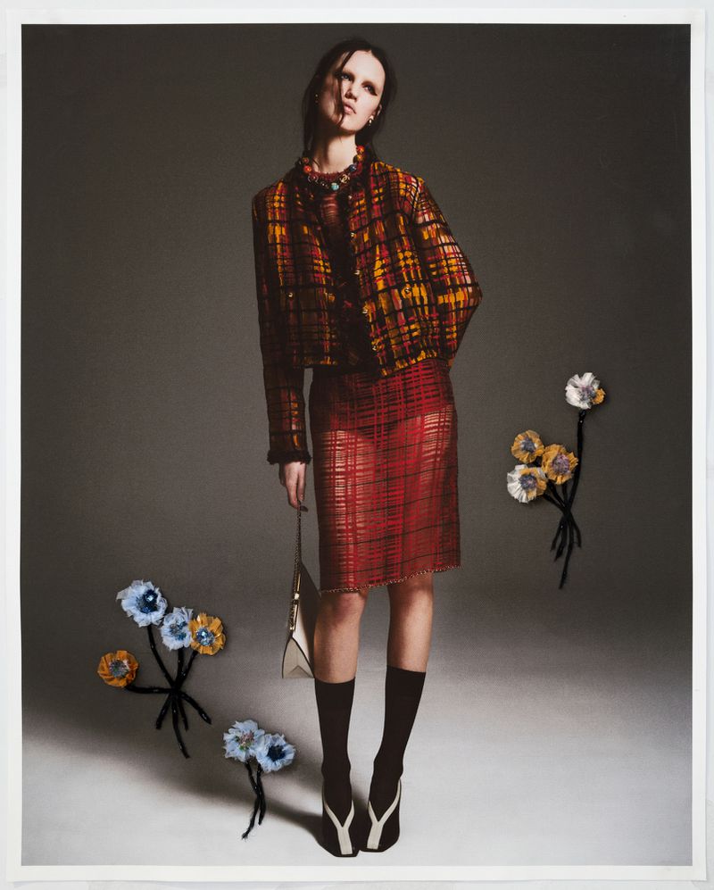 A tall female model poses in a brown plaid jacket and red plaid skirt with a purse, accessorized with floral embellishments on a grey studio backdrop.