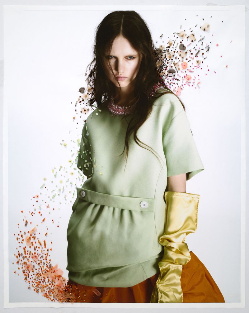 A fashion shot featuring a woman in a green dress with unique embellishments and long gold gloves, standing against a white backdrop.