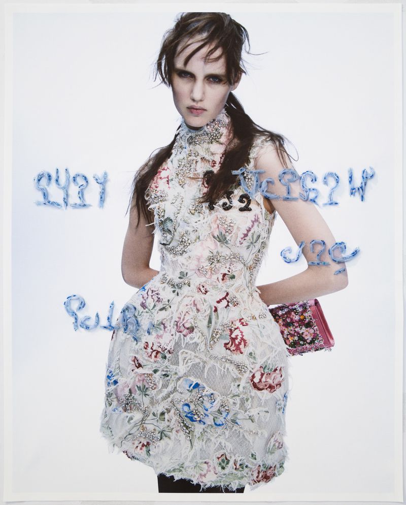 An adult female fashion model poses in a high-necked floral mini dress with embroidery, holding a floral purse on a bright background.