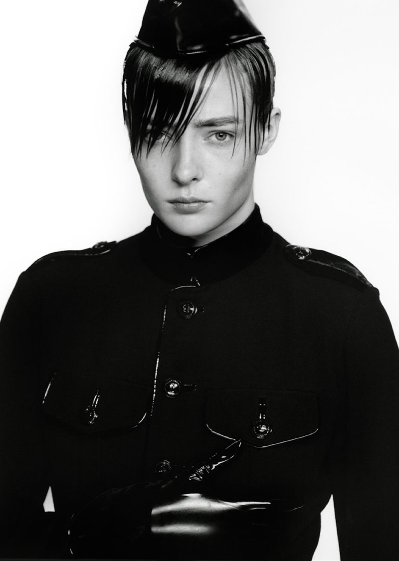Striking portrait of a young adult with dark hair, gazing forward, wearing a dark military-style jacket and cap against a white backdrop.