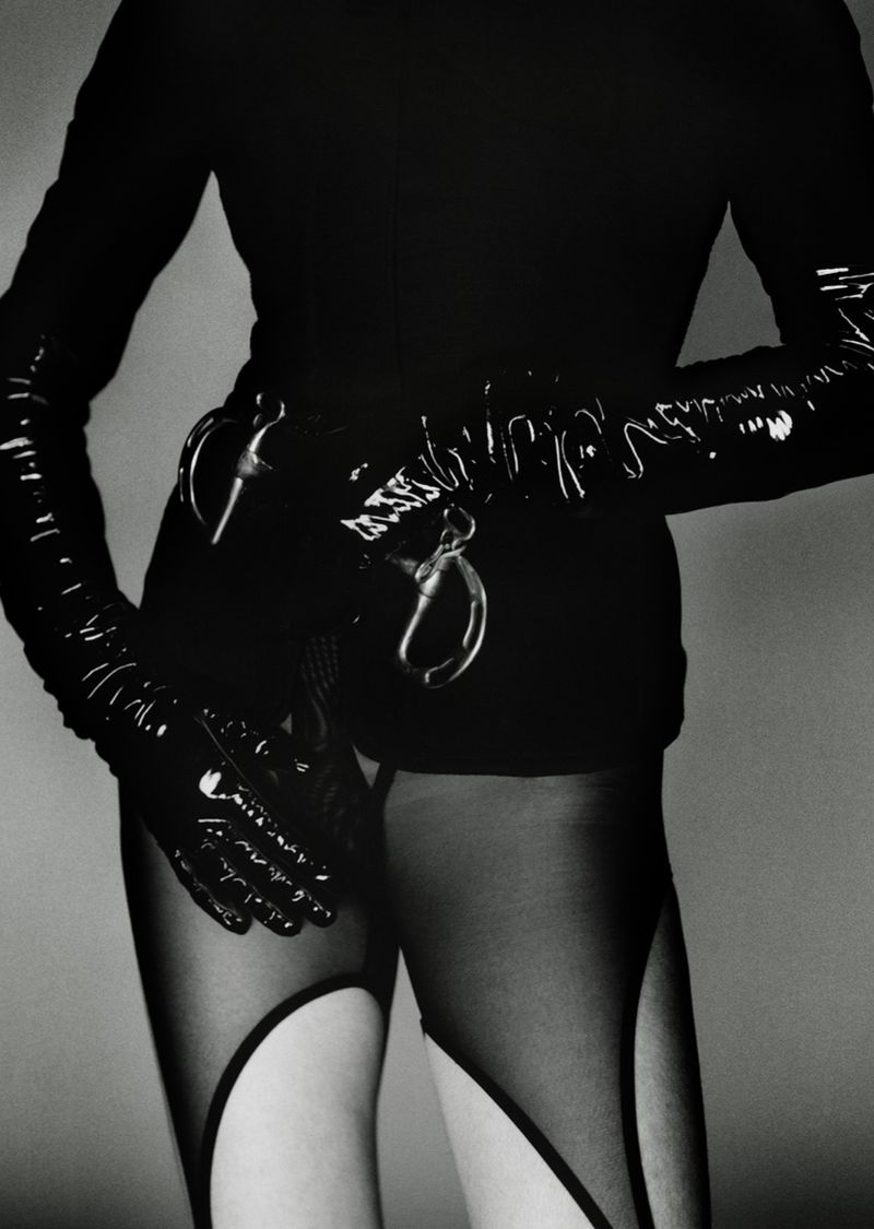 Monochrome image of a person with long, dark gloves holding handcuffs at the waist. The person wears a dark top and leggings.