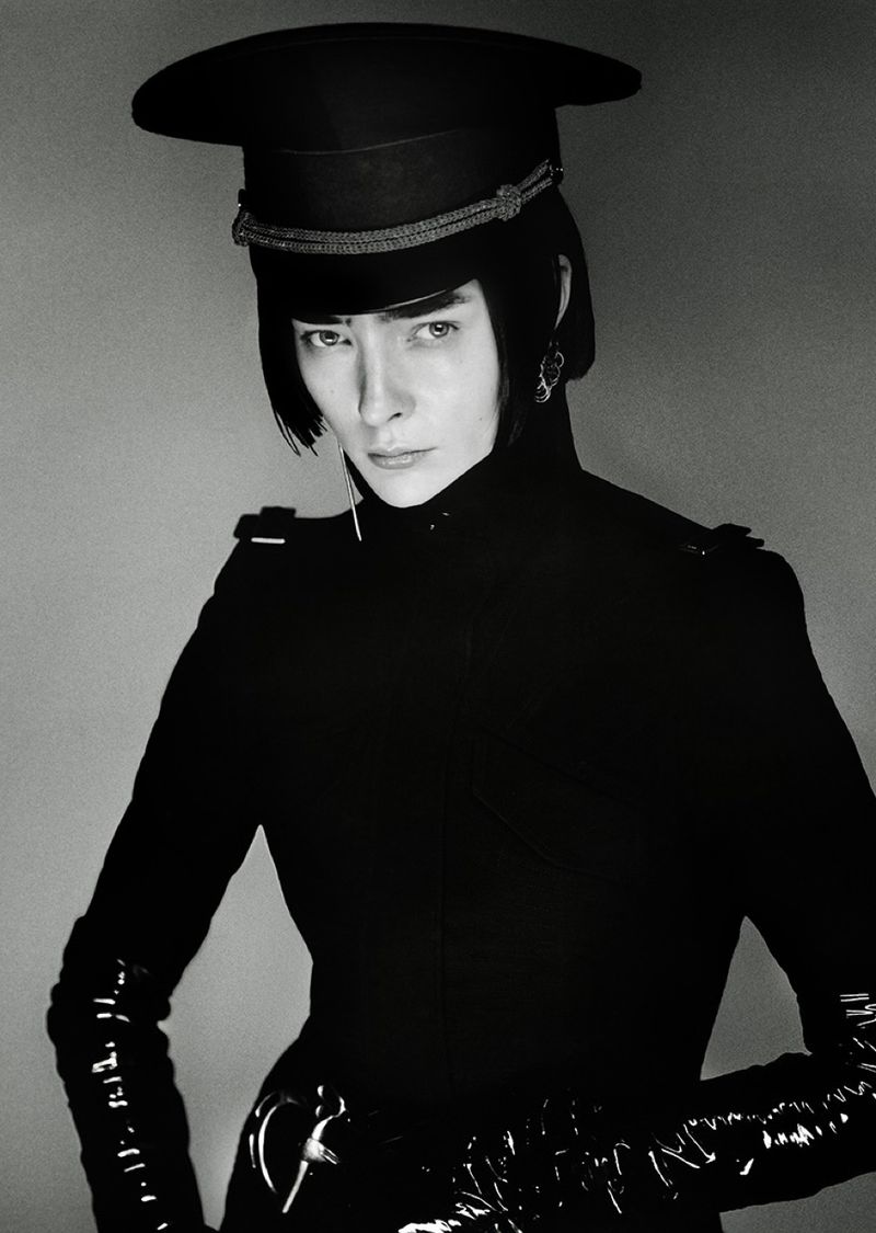 Monochrome portrait of an adult woman in a black military-style hat and jacket with shiny gloves, posing against a grey backdrop.