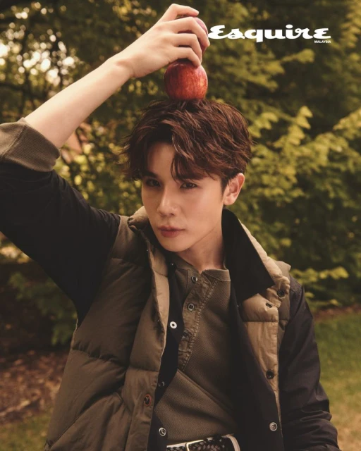 Stylish man poses outdoors with an apple on his head for a magazine cover, wearing a vest and button-down shirt in a natural setting.