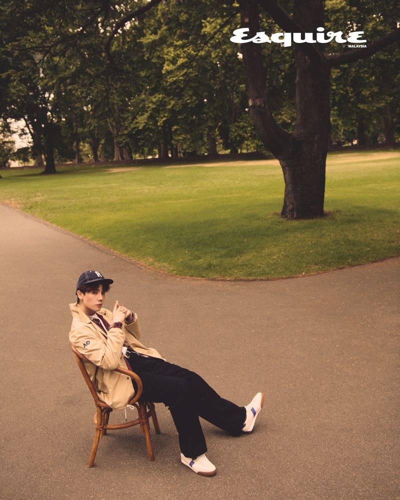 Editorial fashion shot of a young adult man sitting on a chair in a park, wearing a light jacket, dark pants, and a hat.