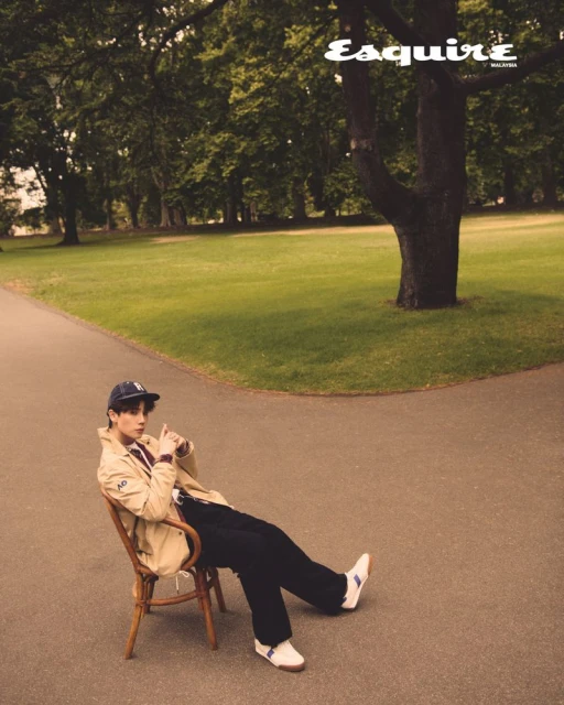 Editorial fashion shot of a young adult man sitting on a chair in a park, wearing a light jacket, dark pants, and a hat.