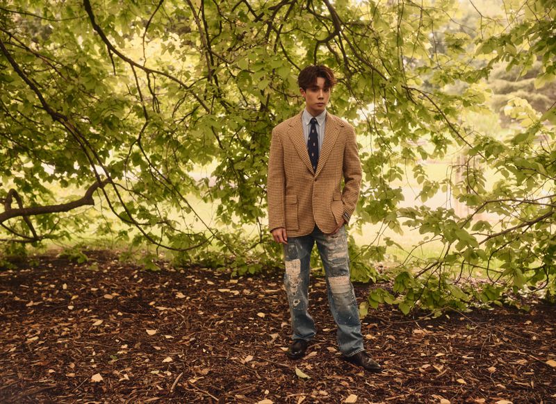 Adult male posing in a tweed jacket and ripped jeans, standing outdoors amidst the lush green trees in a natural setting.