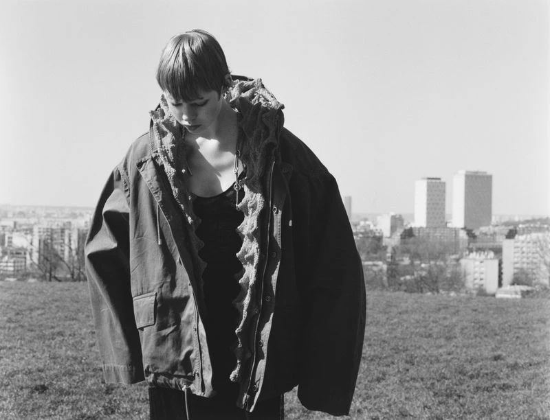 Monochrome portrait of a person in an oversized jacket standing in a grassy field with a cityscape in the background under a bright sky.